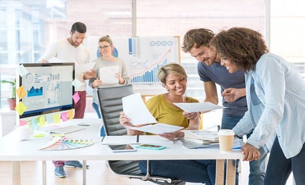 Five diverse people in an office smiling and looking over documents