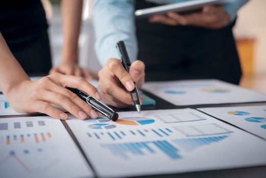 Two people analyzing data charts and graphs on paper spread across a table. One person is pointing at a chart with a pen, while the other is holding a pen and possibly a tablet or notepad. The charts feature various types of data visualizations, including bar graphs and pie charts, suggesting a business or financial context.