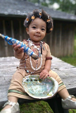 Baby sitting on wooden table outside dressed in traditional native clothes holding an abalone shell and sage bundle