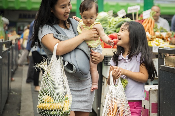 A woman holding a baby and standing with a young girl at an outdoor market, all smiling and carrying mesh bags filled with fresh vegetables.