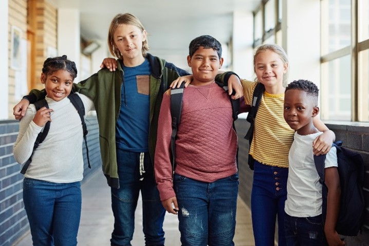 Five children of different genders, ages, and races standing with their arms around each other shoulders smiling at the camera