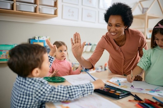 Teacher smiling and high-fiving small child as they paint at a table with two other little kids